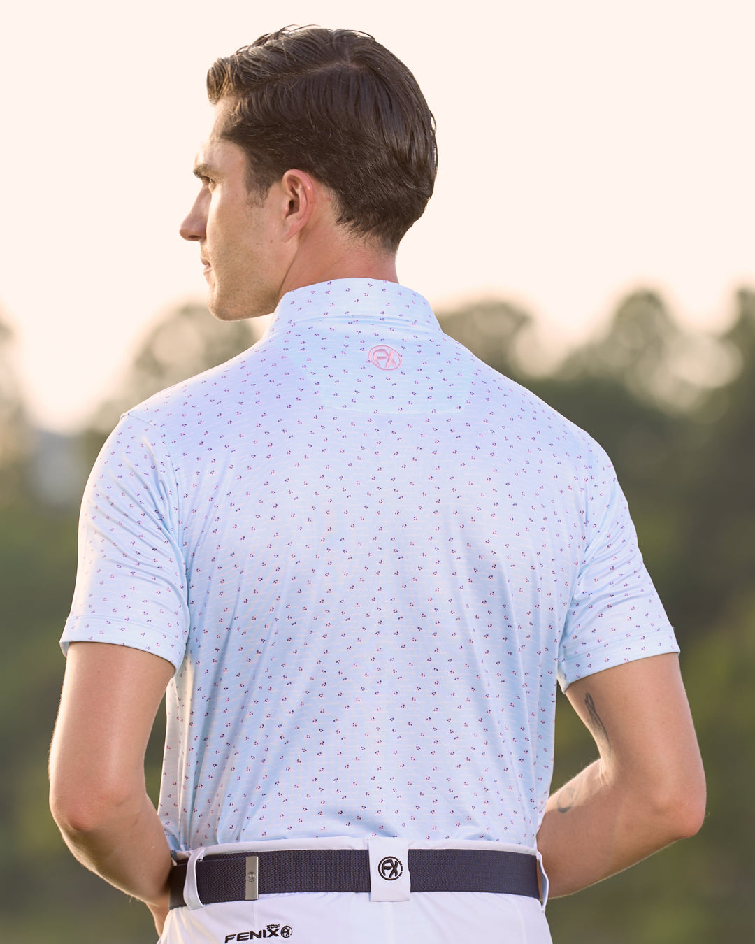 Man in light blue polo shirt and pants standing by a lake with blurred greenery in the background