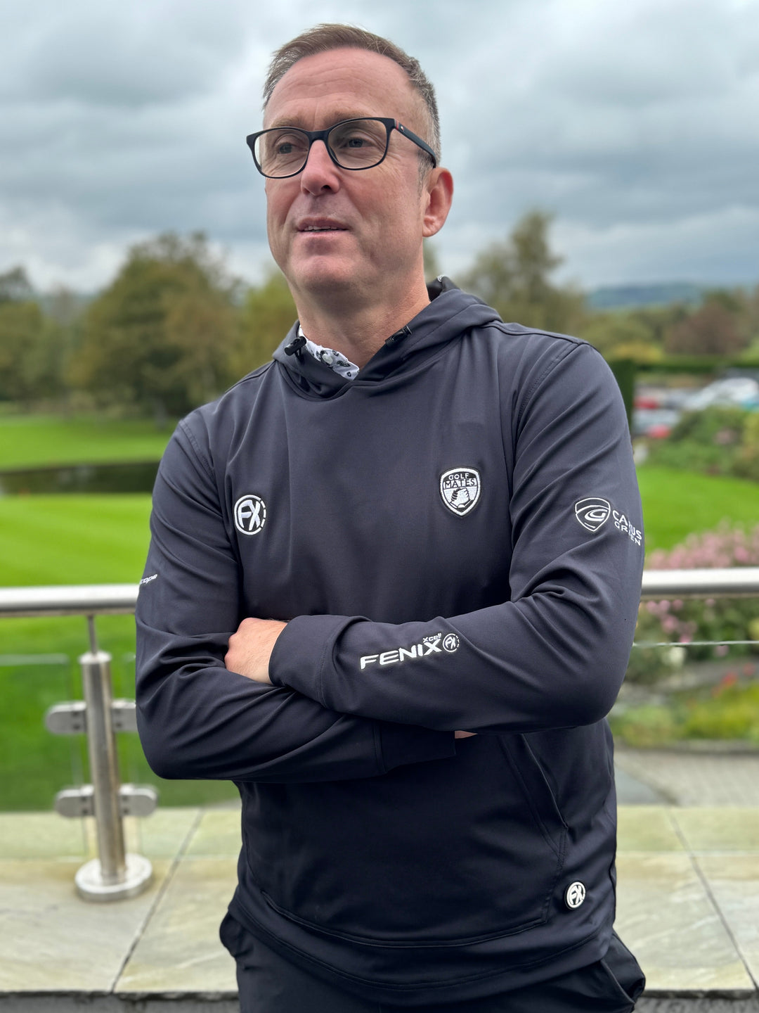 Man wearing a dark jacket with logos standing outdoors on a cloudy day.